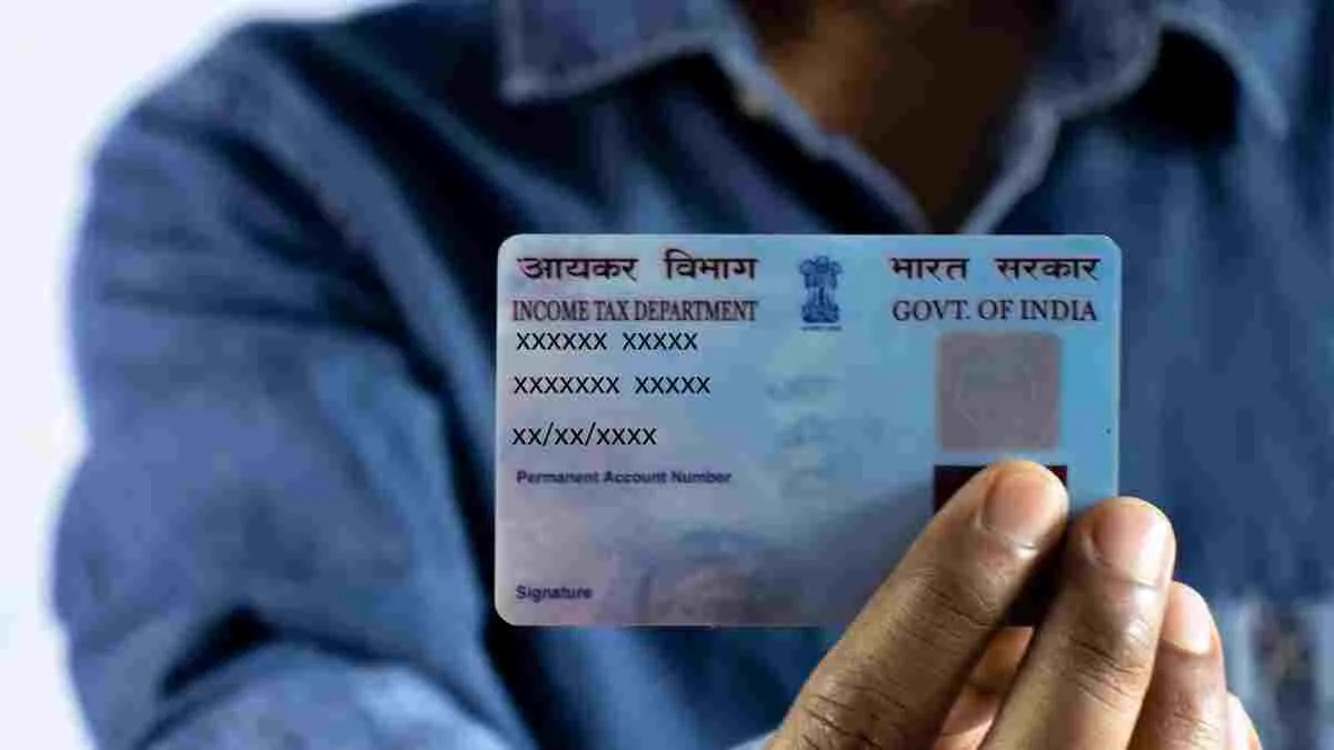 Hand holding an Indian PAN card. The card displays "Income Tax Department" and "Govt. of India" with obscured numbers. Blue shirt in background.